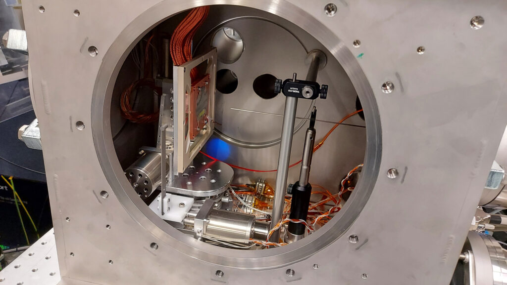 Speziell für das Experiment angefertigte Vakuumkammer: Der runde Wafer – an der linken Seite senkrecht auf einer Kupferoberfläche montiert – ist mit Platin beschichtet und wurde auf -173 °C (100 Kelvin) abgekühlt. Die dünne Stahldüse in der Mitte, die direkt auf den Wafer gerichtet ist, setzte Wasserdampf frei, was zur Bildung von amorphem Eis auf dem Wafer führte. Dieses Eis wurde anschließend durch einen optischen Laser angeregt und mit Röntgenlicht untersucht, das durch das halbverschattete dunkle Loch an der Rückseite der Kammer einfiel. (Foto/©: European XFEL)